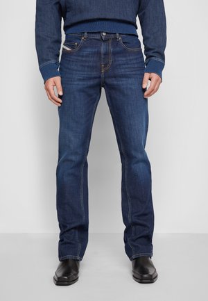 Mann trägt dunkelblaue Jeans mit geradem Bein, blaue Denimjacke und schwarze Lederschuhe, steht vor einem einfarbigen Hintergrund.