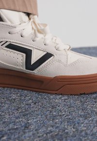 Fehér sneaker, világosbarna gumitalppal, fekete geometrikus logóval, fűzőlyukakkal és mintás felsőrésszel. A fűzők meg vannak kötve és láthatóak.