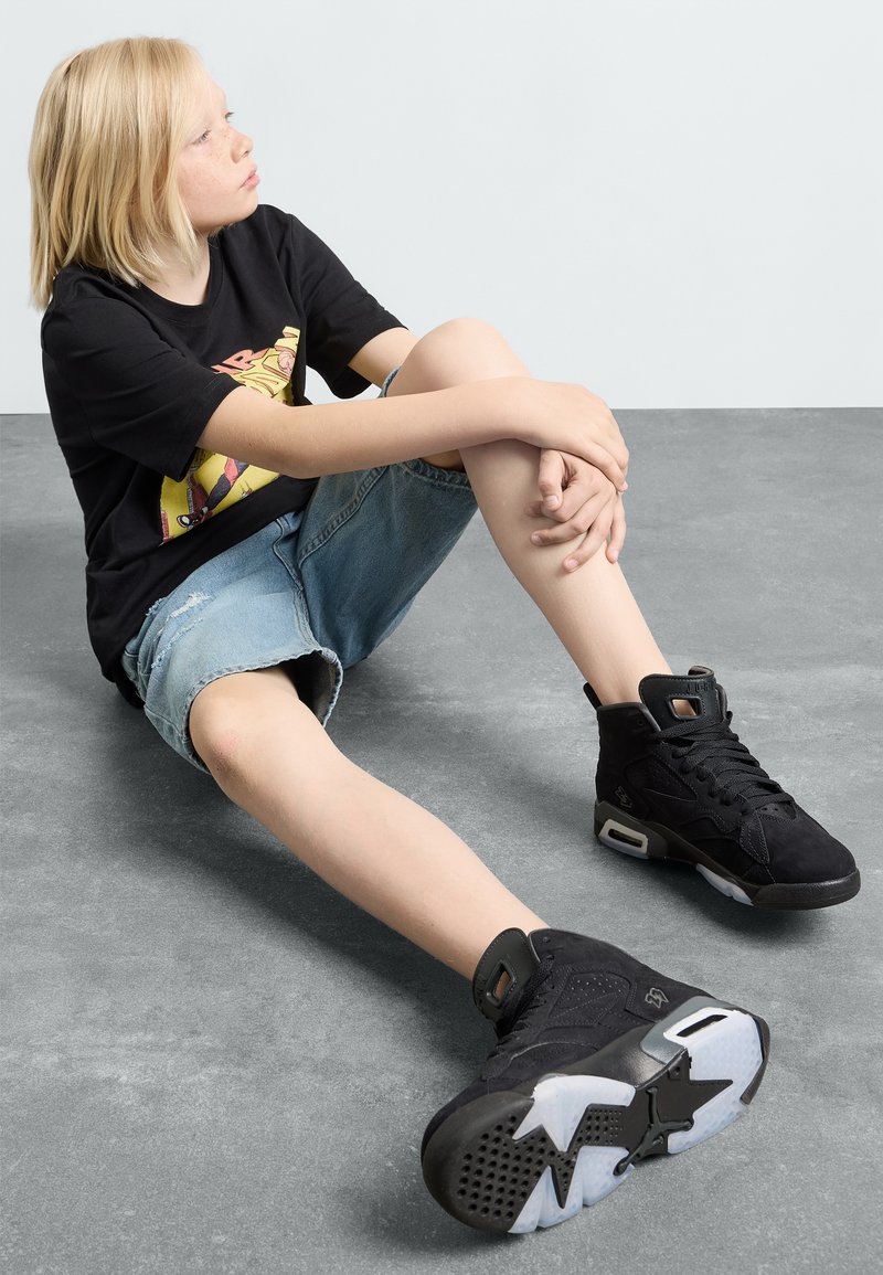 Baskets montantes noires en daim, avec une semelle intermédiaire blanche et une semelle extérieure en caoutchouc. Portées avec un short en jean et un t-shirt graphique noir.