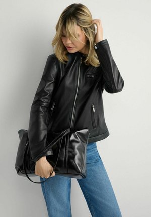 Blondynka w czarnej skórzanej kurtce i niebieskich dżinsach trzyma dużą czarną skórzaną torebkę na jasnym tle.