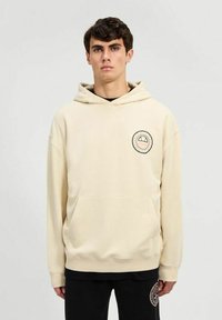 Sudadera color crema hecha de tejido suave con un bolsillo canguro y un logo circular en el pecho. Incluye capucha con cordón.