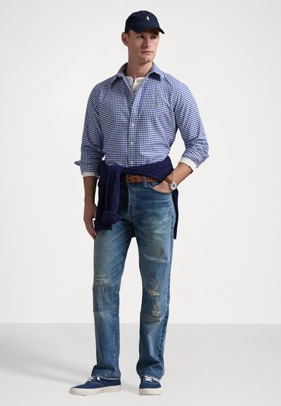 Blauw en wit geruit overhemd met knopen, marineblauwe sweater om de taille, lichtblauwe versleten jeans, marineblauwe sneakers, zilveren horloge.
