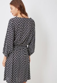 Mujer de perfil que lleva un vestido azul marino con estampado geométrico blanco, mangas abullonadas de tres cuartos y cintura entallada.