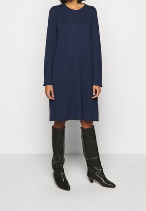 Osoba w granatowej, sięgającej do kolan sukience z długim rękawem, w zestawie z czarnymi, wysokimi na kolano kozakami na obcasie, stojąca na białym tle.