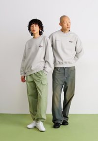 Two models stand side by side wearing light gray sweatshirts with small logos; one wears green cargo pants and white sneakers, the other wears faded jeans and black shoes.