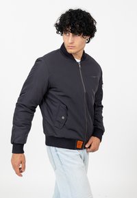 Marineblauwe bomberjack met een rits aan de voorkant, geribbelde manchetten en zoom, zijzakken, en een oranje label aan de linkeronderkant. Soepele textuur.