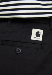 Crne pamučne hlače s prednjim džepom i crnim dugmetom. Bijela kvadratna oznaka ima Carhartt logo u crnoj boji. Iznad je prugasta majica.