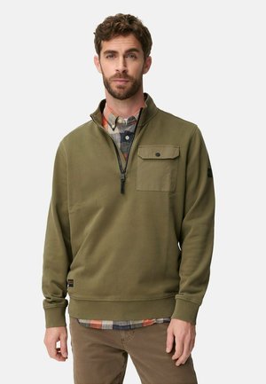 Mann trägt eine olivgrüne Sweatjacke über einem mehrfarbigen karierten Hemd und braune Hose, steht vor einem schlichten Hintergrund.