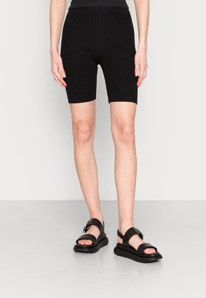 Persona que lleva puestos shorts tipo biker negros con textura acanalada y sandalias negras con punta abierta, suelas gruesas y correas en el tobillo, de pie sobre un suelo gris.