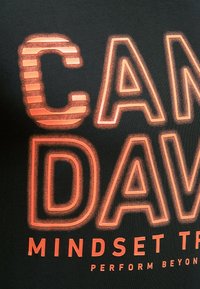 Schwarzes T-Shirt mit auffälligem orangefarbigem Text, der "CAN'DAV" lautet, wobei das "C" eine gestreifte Textur hat. Der Stoff wirkt weich und glatt.