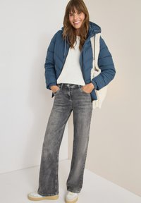 Giacca blu imbottita con cappuccio, maglione bianco e jeans grigi a gamba larga, completati da sneakers color crema e una borsa tote color crema.