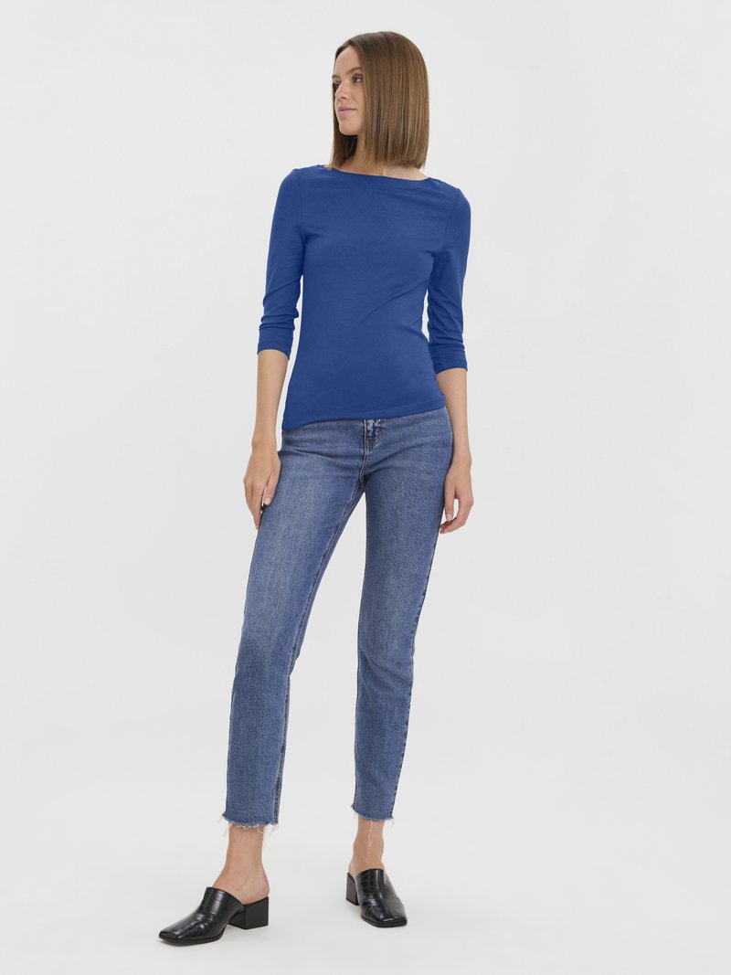 Frau mit glattem, schulterlangem Haar, die ein blaues Oberteil mit Dreiviertelärmeln, verblasste Jeans in Blau mit ausgefransten Saum und schwarze, elegant-absatzlose Slipper trägt.