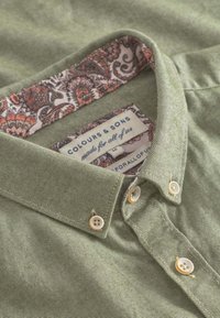 Camicia a righe verdi con un colletto bordato di un motivo floreale, dotata di bottoni bianchi e un'etichetta di stoffa cucita all'interno.