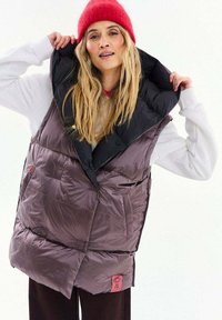 Žena nosi červenú pletenú čiapku, bielu blúzku s dlhým rukávom a veľkú fialovú prešívanú vesta s čiernym golierom, drží golier oboma rukami zdvihnutý.