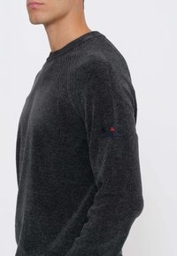 Maglione grigio scuro con motivo a maglia strutturata, caratterizzato da una scollatura a costine e una toppa nera con dettagli rossi sulla manica sinistra.