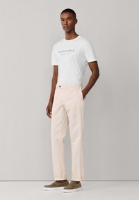 Uomo con indosso una maglietta bianca a maniche corte con scritta, pantaloni chino beige e scarpe senza lacci marroni, in piedi con una mano in tasca.