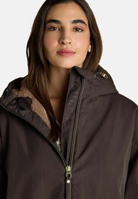 Sieviete ar garām brūnām matēm valkā tumši brūnu jaku ar zippu, kam ir vilnas oderējums un zelta rāvējslēdzēja detaļas uz viendabīga fona.