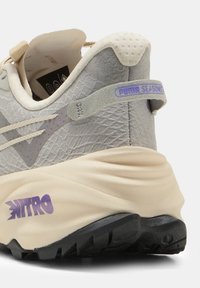 Scarpa sportiva grigia con tomaia testurizzata, suola beige, dettagli viola e il marchio "NITRO" ben in evidenza sulla parte intermedia, dotata di linguetta sul tallone.
