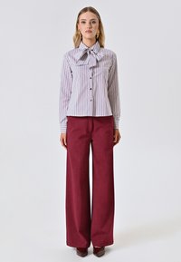 Camicia bianca con strisce verticali bordeaux, colletto a fiocco e frontale con bottoni; abbinata a pantaloni larghi bordeaux e scarpe coordinate.