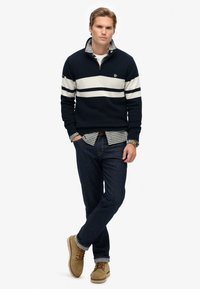 Granatowy sweter z dzianiny z białymi paskami, z zamkiem przy kołnierzu, na koszulę w kratę, ciemne dżinsy i beżowe botki. Swobodny strój z warstwowymi teksturami.