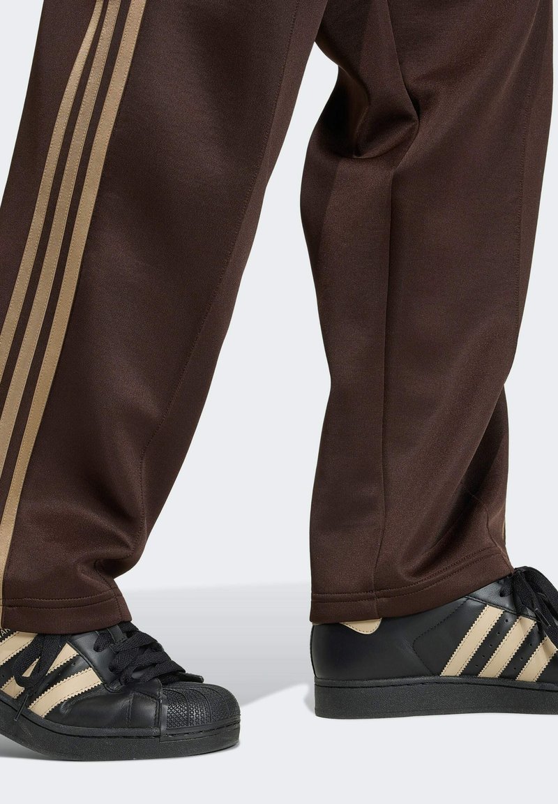 Person trägt braune Trainingshose mit beigen Streifen und schwarze Sneakers mit beigen Streifen, steht auf hellem Hintergrund.