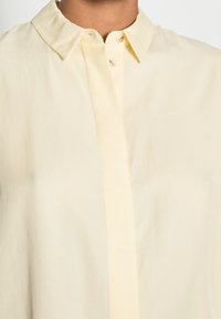 Chemise col jaune clair en tissu lisse ; présente une fermeture à bouton et un design épuré et minimaliste.