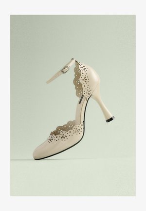 Zapato de tacón alto beige con un diseño de recortes en forma de concha y correa en el tobillo. Presenta un acabado brillante y un tacón con punta negra.