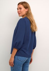 Blusa blu navy con tessuto testurizzato, vestibilità ampia e maniche a sbuffo. Abbinata a jeans in denim aderenti. Vista posteriore mostrata.