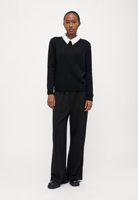 Pull en maille noir avec col en V et chemise blanche à col, accompagné de pantalons larges noirs et de chaussures noires imposantes. Tissu lisse et texturé.