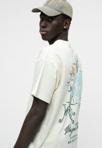 Crème kleurig katoen T-shirt met korte mouwen, voorzien van een gedetailleerde grafische print op de rug in groene en blauwe tinten.