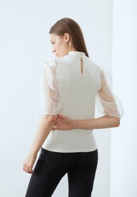 Vit topp med genomskinliga puffärmar och en liten nyckelhålsöppning i ryggen. Matchad med mörk denimjeans. Slät textur och figurnära design.