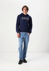 Sweat à capuche bleu marine avec un logo blanc et rouge, associé à un jean bleu clair. Le mannequin porte des chaussures noires, debout sur un fond neutre.