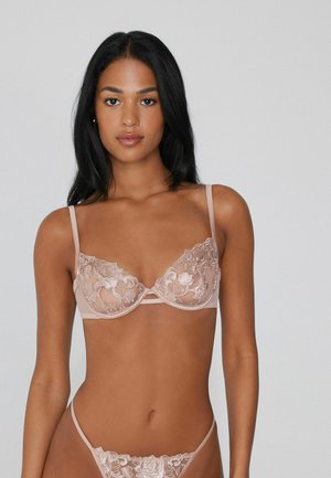 Reggiseno in pizzo di un delicato beige con ricami floreali, supporto con ferretto e spalline regolabili. Include il perizoma trasparente abbinato.