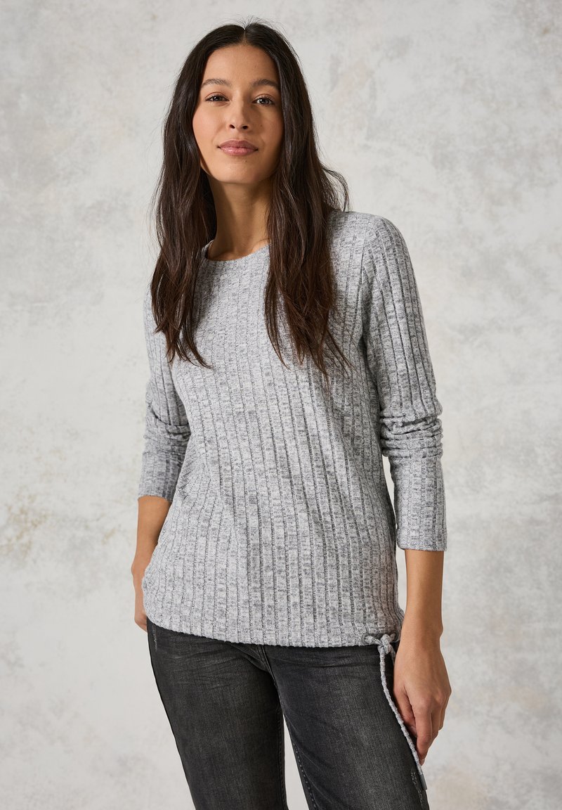 Grijze geribbelde lange mouwen top met zijsnoerdetails, gemaakt van gestructureerde stof. Gecombineerd met donkere jeans. Neutrale achtergrond.