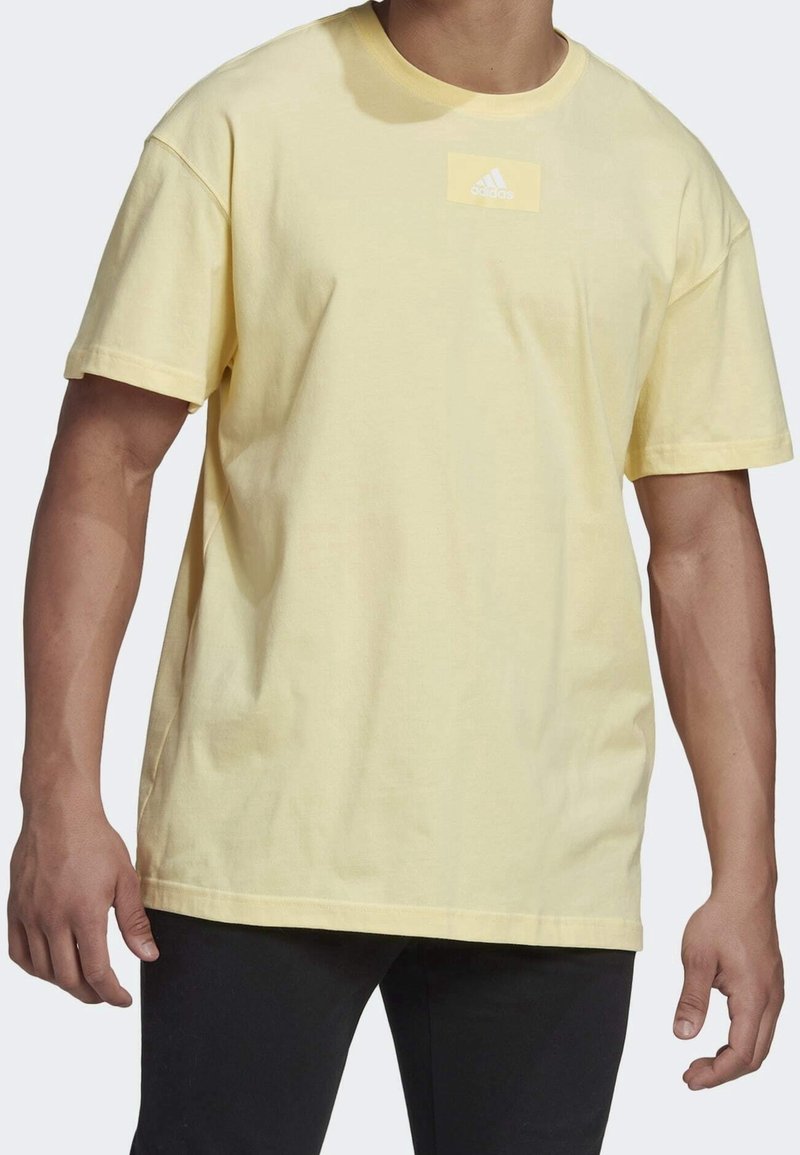 T-shirt di cotone giallo con una vestibilità comoda, maniche corte e una patch rettangolare bianca con tre strisce sul petto.