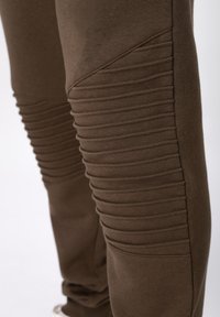 Καφέ φόρμα με ριμπ υφή και λοξές ραφές στα γόνατα, κατασκευασμένη από μαλακό, ελαστικό ύφασμα για άνεση και κινητικότητα.