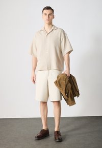 Camisa corta de color beige claro con botones en la parte delantera, combinada con pantalones cortos de color crema. Calza zapatos de cordones marrón oscuro y sostiene una chaqueta de pana en la mano.
