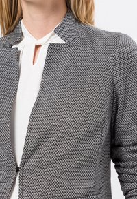 Veste à motif noir et blanc avec une coupe ajustée, un col cranté et une texture lisse. Présente un détail de fermeture subtil à l'avant.