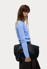 Svart läderhandväska med en mjuk, slapp design, hålls under armen. Personen bär en ljusblå tröja och en mörk kjol.