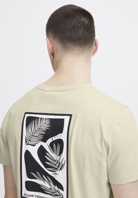 Lys beige t-skjorte med et stort svart-hvitt grafikkmotiv med planteformer på baksiden. Kort ermer og rund hals.