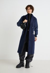 Navy long coat with a double-breasted design, featuring textured fabric and side pockets. Paired with dark pants and black puffy boots.