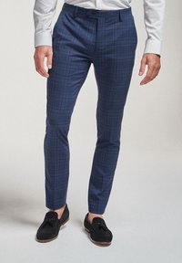 Homme portant un pantalon ajusté à carreaux bleu marine, une chemise blanche habillée et des mocassins noirs en daim avec pampilles, debout sur un fond uni.