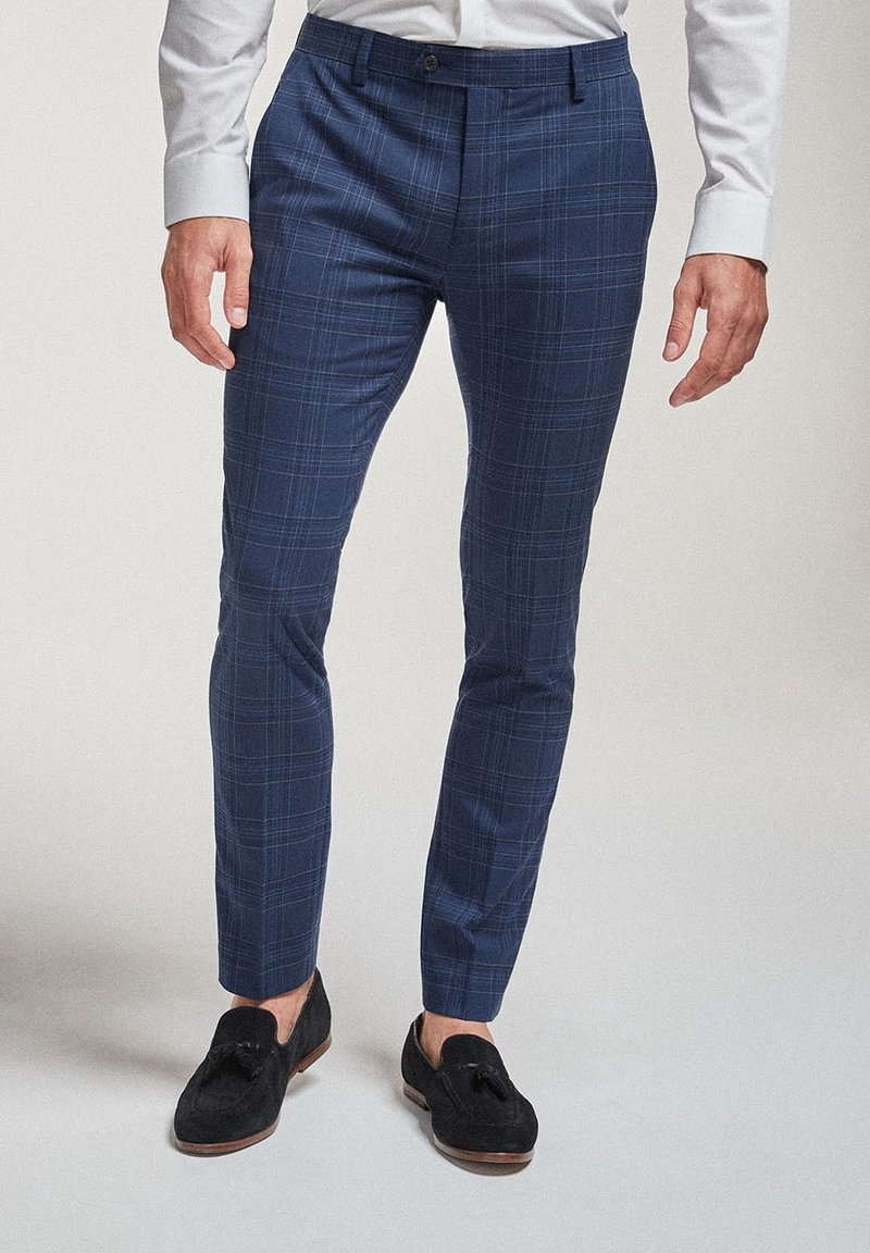 Homme portant un pantalon ajusté à carreaux bleu marine, une chemise blanche habillée et des mocassins noirs en daim avec pampilles, debout sur un fond uni.