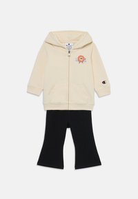 Felpa con zip crema con grafica di leone e testo blu; abbinata a leggings neri svasati. Tessuto in misto cotone, dotata di due tasche frontali.