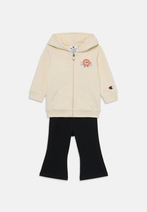 Felpa con zip crema con grafica di leone e testo blu; abbinata a leggings neri svasati. Tessuto in misto cotone, dotata di due tasche frontali.
