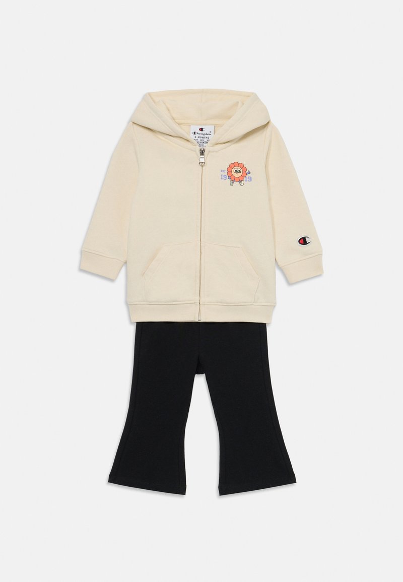 Felpa con zip crema con grafica di leone e testo blu; abbinata a leggings neri svasati. Tessuto in misto cotone, dotata di due tasche frontali.