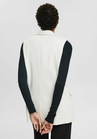 Weißer ärmelloser Blazer mit strukturiertem Design, ausgestattet mit einem Rückenschlitz, kombiniert mit langen schwarzen Rippenärmeln.