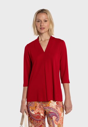 Czerwona bluzka z dekoltem w serek i rękawami do łokcia, wykonana z miękkiego, lekkiego materiału o minimalistycznym designie, zestawiona z wzorzystymi spodniami.