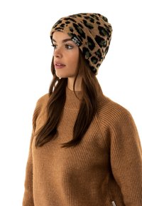 Berretto a maglia leopardato beige con una texture morbida, caratterizzato da un risvolto, indossato con un maglione a coste color cammello. Etichetta notevole sul berretto.