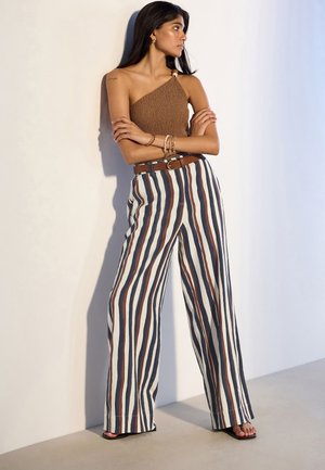 Mujer de pie con los brazos cruzados, vistiendo un top marrón de un solo hombro y pantalones anchos de rayas de talle alto con sandalias frente a una pared blanca.